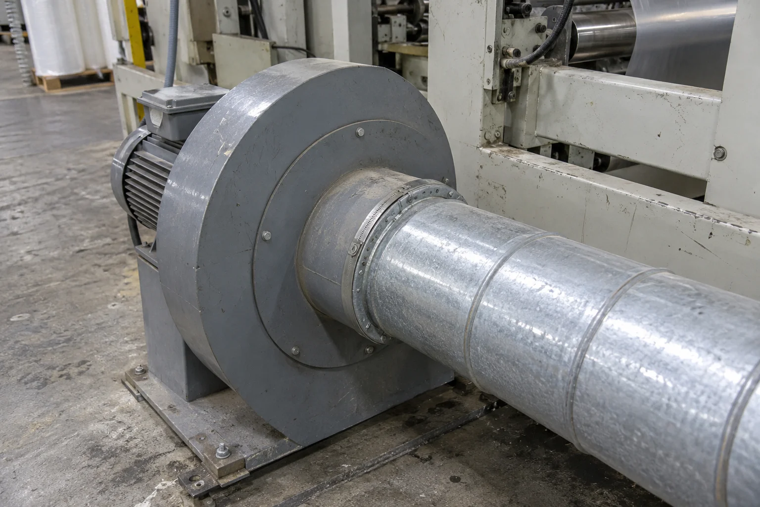 A grey blower housing connects to a round metal duct beside a machine frame.