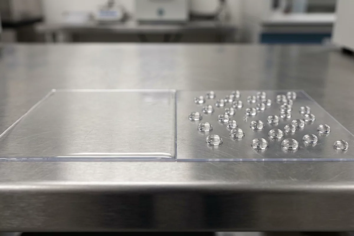 A macro photograph comparing two liquid test swatches on a plastic substrate, contrasting uniform wetting against beading droplets.